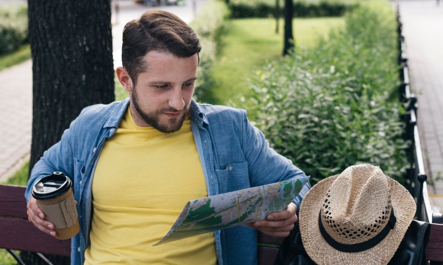 A man in a yellow shirt reading a map on a park bench with a coffee